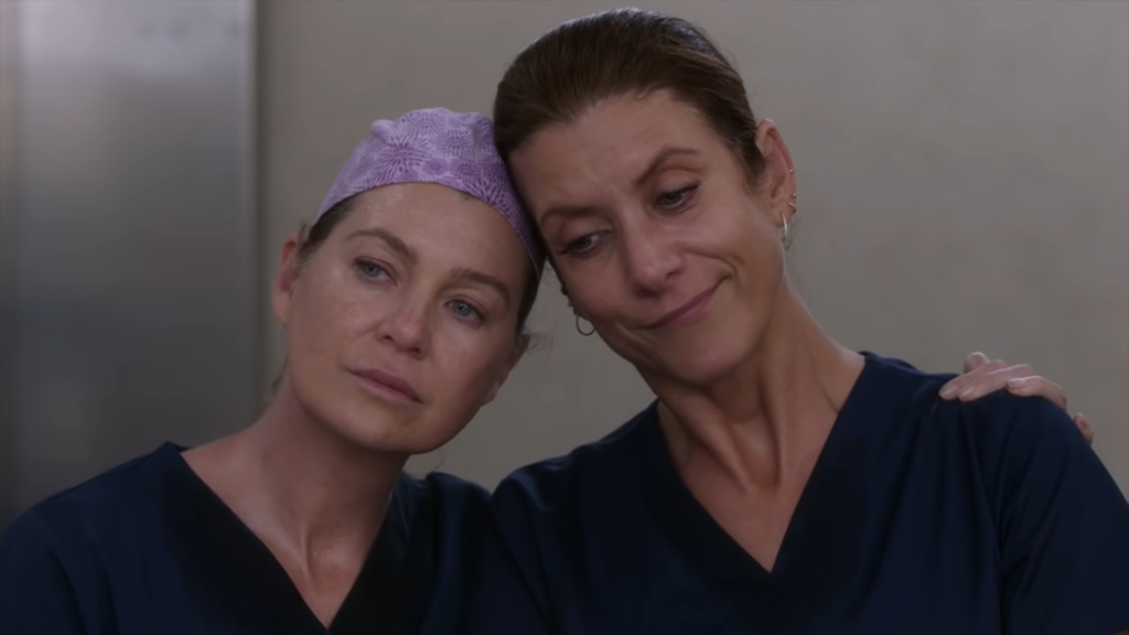 Meredith (played by Ellen Pompeo) wearing her scrub cap with her head leaning against Addison's (played by Kate Walsh), who's no longer wearing her scrub cap, in the elevator