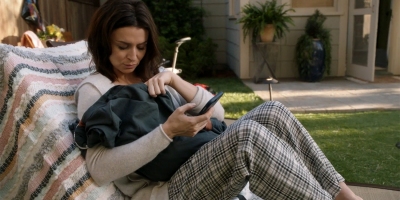 Still of Amelia (played by Caterina Scorsone) holding a baby Scout under a blanket while holding her phone in her other hand while sitting in a piece of outdoor furniture from Grey's Anatomy 17x02